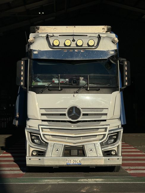 A front view of a large Mercedes-Benz delivery truck parked inside a warehouse or loading dock, with a dark interior background. The truck is predominantly white with a silver Mercedes-Benz emblem on