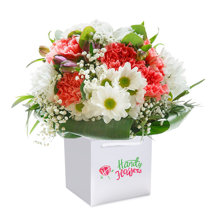 A lush and vibrant bouquet, lovingly composed by Queen's Park Florist, is showcased upright within a crisp white square gift bag, set before a spotless white background that highlights every exquisite detail. The arrangement pulses with the warmth of coral, peach, and deep red carnations, their ruffled, soft petals layering together to create a delightfully textured focal point-evocative of springtime strolls through Queen's Park Gardens. Here and there, pristine white daisy-like chrysanthemums with lively green-yellow centers bring refreshing brightness and an element of playful contrast. Wisps of baby's breath add a light, cloud-like softness beneath the bolder blooms, while small, purple-tinged buds peeking subtly from the side lend intrigue and the anticipation of unfolding beauty. Encircling the gathering, glossy deep green foliage creates a natural collar, reminiscent of the thriving greenery in the park itself. The bouquet's rounded, abundant shape is both classic and contemporary, making it ideal for birthdays, anniversaries, or as a thoughtful thank-you delivered to a friendly neighbour on Harvist Road. Atop the clean, modern bag, sturdy ribbon handles suggest an arrangement designed for effortless local gifting. This bouquet's refreshing palette and balanced design evoke a sense of genuine care, speaking to the spirit of Queen's Park-where community and craft always bloom together.