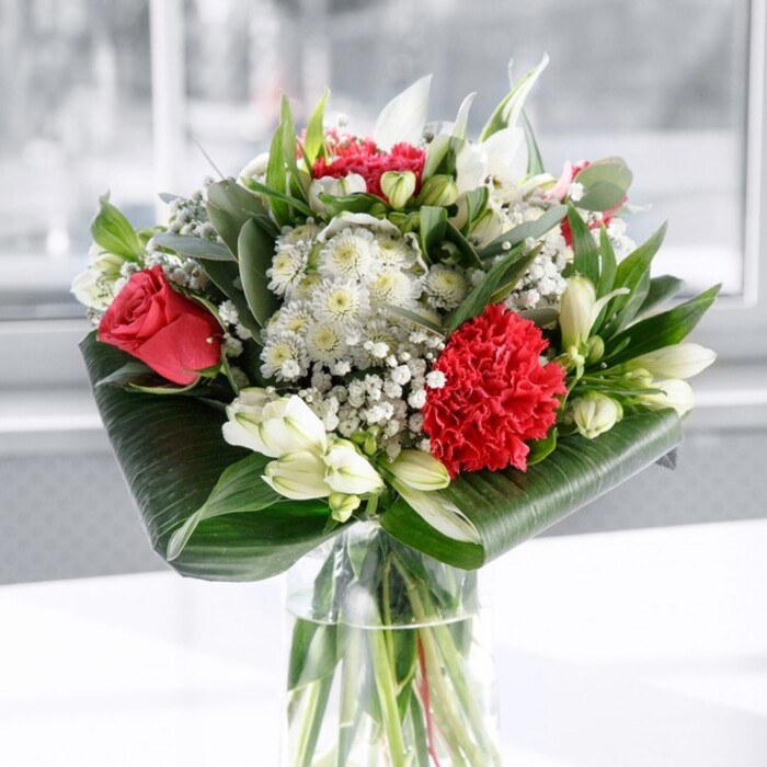 A sophisticated red and white floral arrangement stands in a clear cylindrical glass vase, poised as though ready to grace a dining table in a Queen's Park townhouse. The transparent vase showcases the straight, carefully trimmed green stems beneath the waterline, each stem arranged with deliberate precision, suggesting the practiced hands of an experienced florist. Above the glass, the bouquet swells into a lush dome of colour and texture. Deep red roses dominate the composition, their velvety petals curling open in elegant spirals, each bloom suggesting timeless romance. At the base, a large, intricately layered red carnation offers a ruffled, almost sculptural focal point. The intensity of the reds is tempered by a scattering of white and cream flowers: white alstroemeria with narrow, speckled petals; rounded white chrysanthemums and daisy-like blooms; and several sleek, closed buds of white lilies that stand upright, promising beauty yet to unfold. Between these larger flowers, clusters of tiny white gypsophila create a misty veil, softening transitions and adding an ethereal glow. Surrounding the blossoms, greenery plays a key structural role. Broad, glossy dark green leaves curve around the base like a tailored collar, while smaller, eucalyptus-style sprigs and pointed green leaves rise through the flowers, introducing subtle variations in tone and shape. The arrangement rests on a spotless white surface against a softly blurred, bright background, recalling the gentle daylight that filters through windows along streets near Queen's Park itself. The lighting is even and calm, allowing the red blooms to look rich without harshness and the white petals to appear clean and serene. The overall mood is warm, polished, and welcoming-perfect as a centrepiece for an anniversary meal, a thoughtful gift delivered locally, or a refined gesture of gratitude to someone in the Queen's Park community.